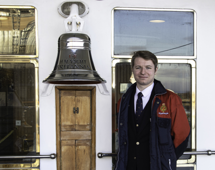 Male Visitor Assistant stands next to Britannia's Bell. 