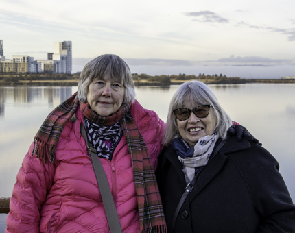 Two women visitors wearing winter coats stand on Britannia's outside deck in front of a view of Leith's waterfront.