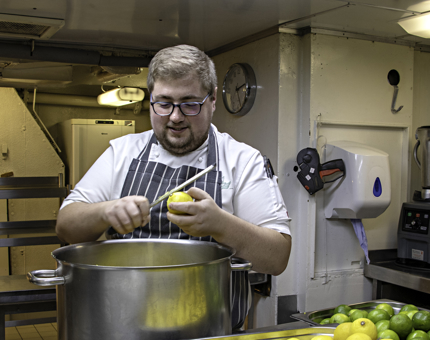 Chef zests a lemon over a large serving pan in the onboard Galley. 