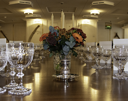 The State Drawing Room table laid for a dinner event with drinks glasses, menus cards and a vase or rich red and orange autumnal flowers. 