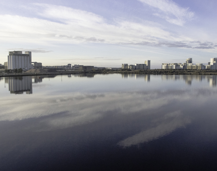 A panoramic view across the waterfront at Leith with tall buildings in the background. There is a blue sky and clouds and the reflection of the sky is mirrored in the water. 