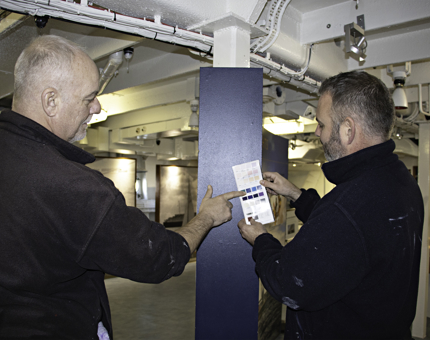 Two Maintenance men hold a paint swatch chart up against a blue panel to match the colour. 