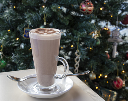 A tall glass of hot chocolate with a chocolate snowflake stencil on top. A lit Christmas tree is in the background. 