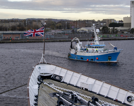 The bow of Britannia is in the foreground with a flag fluttering in the wind. A Marine Research Vessel boat is in the background. 