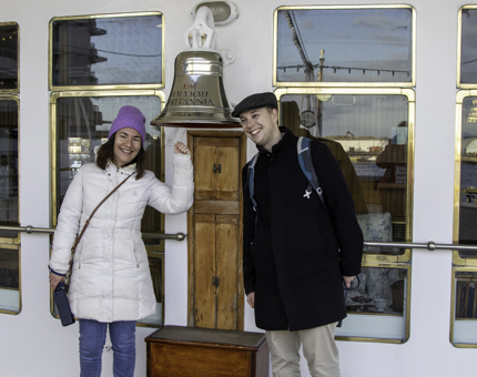 Woman rings Britannia's bell and stands next to another smiling visitor on the Verandah Deck. 
