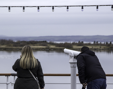 Two people standing on the Verandah Deck. One person is looking through the telescope while the other person looks out across the water.