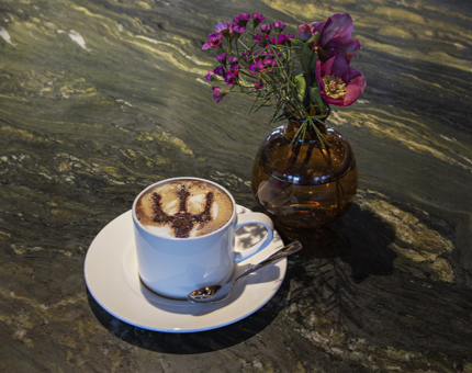 A cappuccino with a Fingal trident stencil on top and a small vase of flowers on table. 
