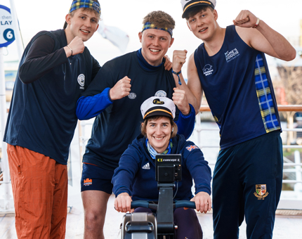 The Weir brothers stand behind Dame Katherine Grainger who sits on a rowing machine. 