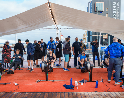 On the Verandah Deck where four team members are rowing on the machines. Team members stand behind cheering them on. 