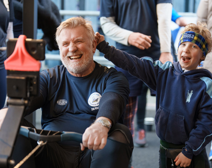 Atlantic rower John Davidson laughing as he rows on a machine with his son standing beside him.
