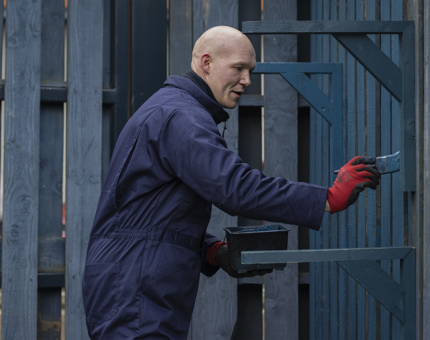 A man holding a paintbrush paints a fence blue. 