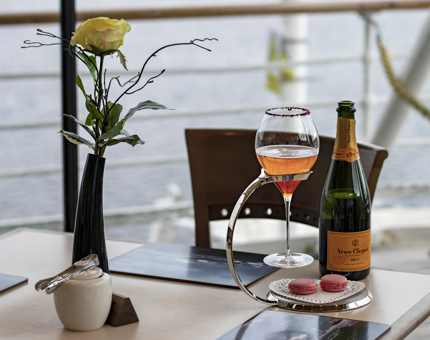 A bottle of champagne, a cocktail in a glass and two pink macrons on a plate with a yellow rose in a vase in the Royal Deck Tea Room. 
