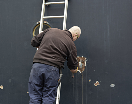 A member of the Maintenance team standing on a ladder at Britannia's hull as he removes blown filler. 