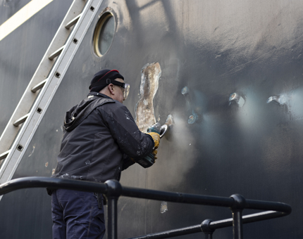 A team member from Maintenance standing next to a ladder holds a tool to clean the steelwork. 