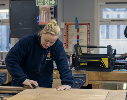 A woman member of the Maintenance team holding a sanding block as she works on a door in the workshop. 