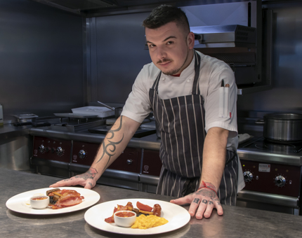 A chef standing in the Galley with two plates in front of him containing Scottish breakfasts. 