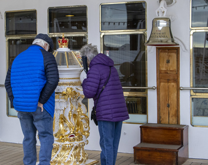 A couple including a man and a woman listen to the audio guided tour handset while looking at the tall white and gold decorative binnacle compass. 