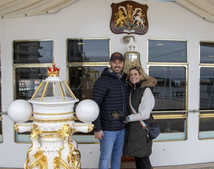 A man and woman couple wearing outdoor coats standing smiling as they pose on Britannia's Verandah Deck. 