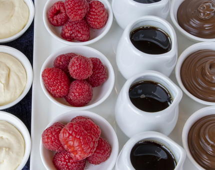 A close-up of small dishes of pancake ingredients including whipped cream, raspberries, syrup and chocolate. 