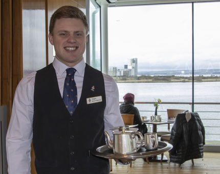 A Waiter standing smiling waiting to deliver a tray of drinks including a teapot, milk jug, cup and saucer in the Royal Deck Tea Room. 