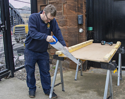 A Maintenance team member standing outside the workshop at Fingal sawing a length of wood. 