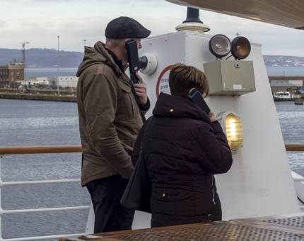 A man and woman visitors stand with their backs to the camera outside on deck listening to audio guide handsets. 