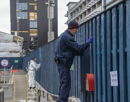 Two Facilities Officers stand on ladders painting Britannia's compound fence blue. One man wears white overalls and the other wears a blue fleece and trousers. 