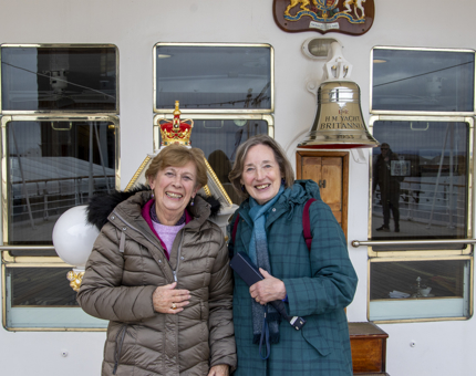 Two lady visitors wearing winter coats standing next to Britannia's shiny bell on deck. The lady on the right is holding an audio guide handset. 