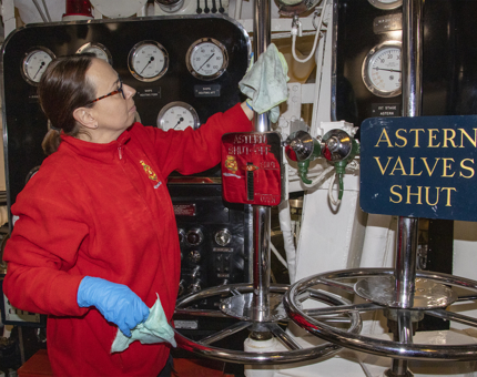 A Housekeeper polishing chrome tubes in the Engine Room. There are several wall mounted dials in the background. 