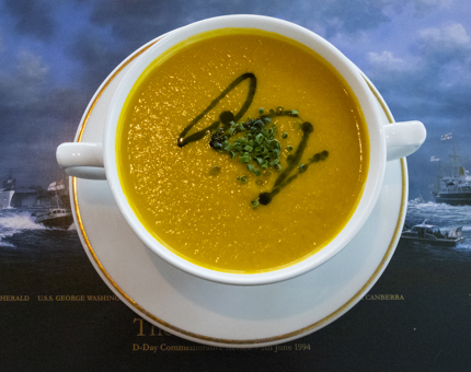 An aerial view of a bowl of sweet potato soup in a bowl sitting on a saucer on a Britannia placemat. 