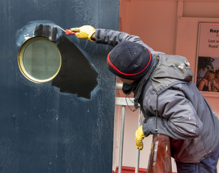 A man from the Maintenance team leaning over a railing holding a roller brush applying undercoat around the outside of a porthole.