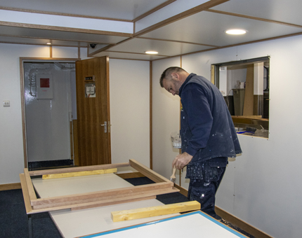 A member of the Maintenance team holding a paintbrush as he varnishes a wooden hatch surround. 