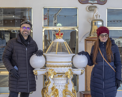 A man and a woman wearing winter coats and sunglasses stand either side of the compass binnacle outside the Sun Lounge. 