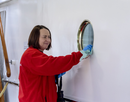 A Housekeeper woman polishing the metal surround on a porthole smiling.