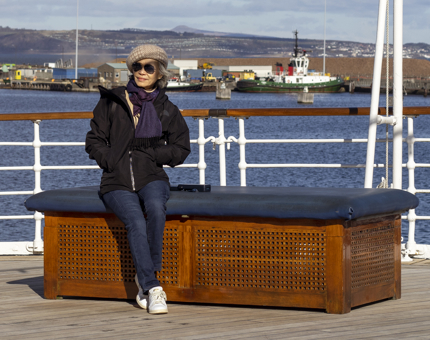 A visitor basking in the sunshine wearing sunglasses sitting on a bench with her hands in her pockets. 