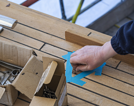 A man preparing an area of wood to fit stanchion pads. There is blue tape marking out the area and small boxes of screws and bolts. 