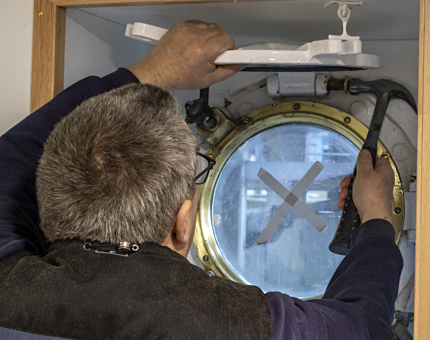 A member of the Maintenance team fitting the deadlight on a porthole. He uses a hammer. 