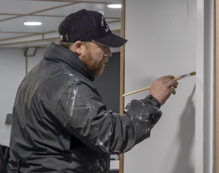 A Maintenance man wearing a baseball cap holding a paintbrush as he touches up the door standard in the former Junior Rates Dining Hall.