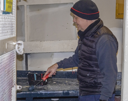 A member of the Maintenance team holding a roller paintbrush as he paints the bench in the aft deck store. 