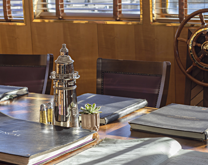 Inside the Bridge at Fingal, the table is laid with large old leather bound books, and a silver lighthouse shaped cocktail shaker. The ship's wheel can be seen in the background. 