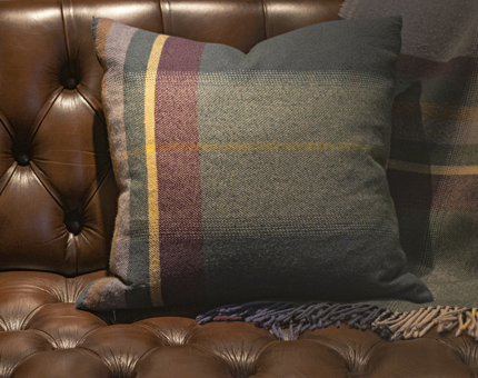 A tartan square cushion in brown, green, yellow, with a throw in the same design sitting on a brown leather Chesterfield sofa. 