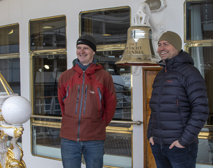 Two visitors wearing outdoors jackets are smiling as they stand next to The Royal Yacht Britannia's Bell. 