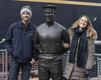 A man and a woman pose next to the bronze statue of former Yottie, Norrie, within the Britannia enclosure. 