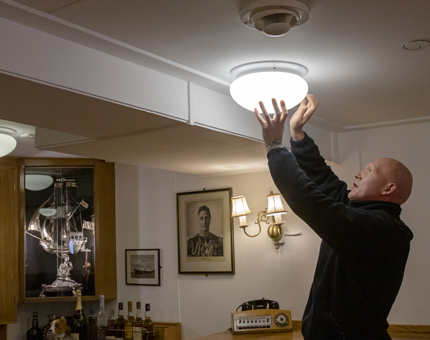 A man from the Facilities team changes a lightbulb from a ceiling lightshade in the Wardroom Anteroom. 