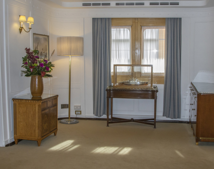 The interior view of the Drawing Room Anteroom on board Britannia. There is a silver model of Britannia in a case, and sideboards with a large vase of flowers. 