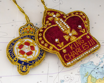 Two embroidered hanging decorations, one the Britannia crest and the other a red and gold crown-shaped decoration embroidered with 'King Charles III'.