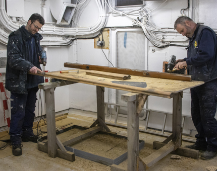 Two Maintenance men holding tools strip back the varnish on handrails.