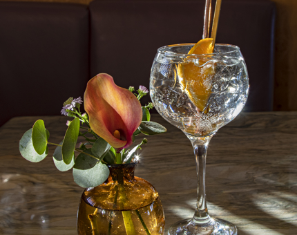 A glass containing a gin and tonic with a slice of orange sitting next to a short amber coloured vase containing an orange lily, purple daisies and stem of eucalpytus. 