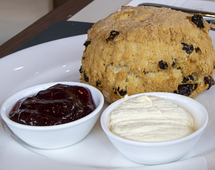 A large fruit scone on a plate with small dishes of clotted cream and jam. 