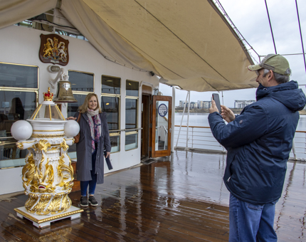 A man taking a photograph of a woman posing next to Britannia's binnacle and in front of the Bell. 